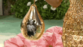 Met Gala 2024 gif. Camera pans up on Sabrina Harrison. She's holding a golden droplet five times the size of her hand that she dangles and sways back and forth. The camera pans up as she turns her face to show the gold bedazzled fascinator resembling a clock face fixed at an angle on her head.