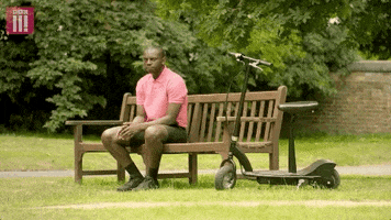 TV gif. Kayode Ewumi as Kazim in Enterprise sits on a bench next to an electric scooter and waves stoically.