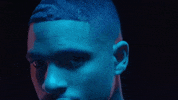 Music video gif. Arin Ray in A Seat music video is washed over in blue and orange light. He moves around, locking eyes with us as he moves his head around.