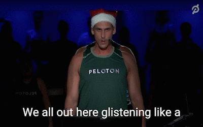 Ad gif. A Peloton ad of a muscly man in a Peloton tank top with a headset microphone and a Santa hat on says, “We all out here glistening like a beautiful Christmas ham right now.”