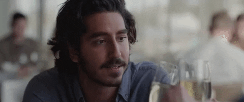 Movie gif. Dev Patel as Saroo in "Lion" clinks champagne glasses with others off-screen and smiles.