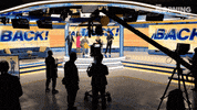 Reality TV gif. We are behind the scenes on the set of The Morning Show as the crew films the anchors cheering up front while blue and yellow streamers fly in the air. Displayed upon the screens behind them, large blue text reads, "Alex is back!"