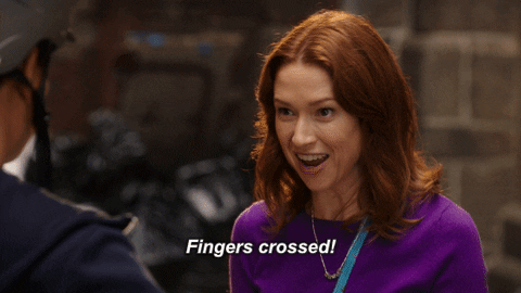 TV gif. Ellie Kemper as Kimmy in the Unbreakable Kimmy Schmidt crosses her fingers tightly on both her faces and has an excited, optimistic grin on her face. She says, “Fingers crossed!”