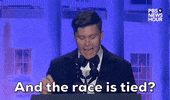 Video gif. Saturday Night Live's Colin Jost stands at a podium at the 2024 White House Correspondents' Dinner as incredulously yells "And the race is tied?"