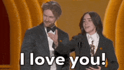 Oscars 2024 gif. Billie Eilish and Finneas O'Connell win Best Song. With her hand, Eilish gestures towards someone in the crowd and says passionately, "I love you!" O'Connell smiles in agreement. 