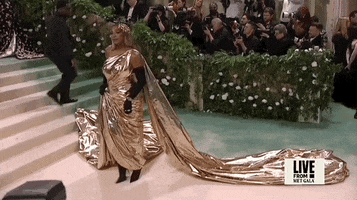 Met Gala 2024 gif. Serena Williams, dressed in gold, moving to walk away is limited by her dress, and covers by striking another pose as assistants adjust her train.
