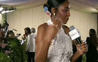 Met Gala 2024 gif. Tilt down on Ayo Edibiri's unfussy Loewe gown of 3D cutout flowers that start white and fade into color as she speaks into a Vogue microphone with gracefully animated gestures.