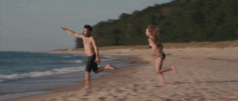 Movie gif. On a beach, Olivia Wilde as Kate in Drinking Buddies gallops after Jake Johnson as Kyle, who trips and falls once he reaches the water.
