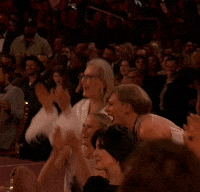 TV gif. Singer Taylor Swift, actress Meryl Streep and producer Mark Ronson stand up to cheer the stage at the 2024 Grammy Awards. Streep and Swift look joyous as they stand up from the audience to cheer, with Streep enthusiastically clapping while Swift excitedly rocks back and forth with a fist up to her nose. Ronson follows suit and is seen in the back standing up and applauding.