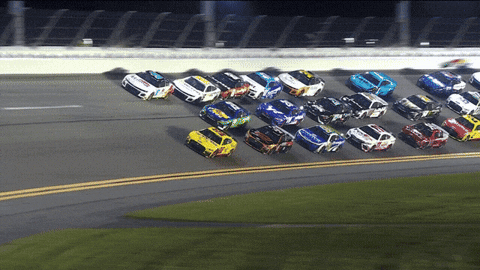 Video gif. Dozens of NASCAR race cars speed around the corner of a track clustered closely together in three long lines. 