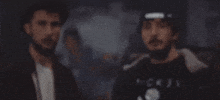 Video gif. Two men stare at us very seriously. A fire then comes into view in front of them.