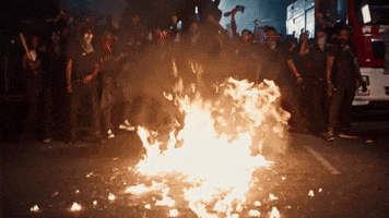 Video gif. Fire burns on the ground and the crowd around it cheers.