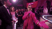 Oscars 2024 GIF. Ryan Gosling, dressed in a Barbie pink tuxedo and sunglasses, reaches out and grabs the disembodied hand of the camera man, holds it, kisses it, then leads him up onto the stage.