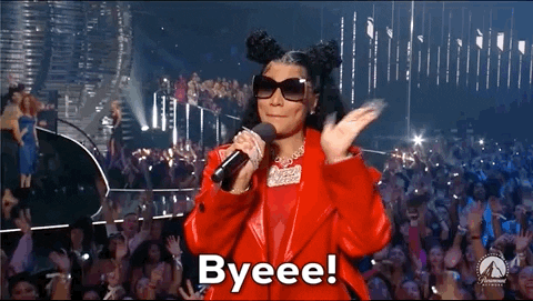 Video gif. Nicki Minaj at the 2023 VMAs, wearing dark oversized sunglasses and a red leather jacket with her hair in half-up-half-down high buns, holding a microphone while smiling and waving at the crowd. Text, "Byeeee!'