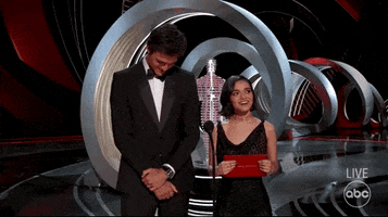 Celebrity gif.  Jacob Elordi and Rachel Zegler are giving an award at the Oscars. Jacob is smiling with his head down and hands crossed and Rachel gives a big smile and raised fist of excitement.