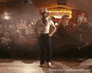 Video gif. A cowboy with a beard starts doing the line dance while the crowd around him watches and claps along. He finds his groove as the spotlight hits him from above.