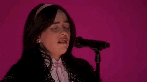 Oscars 2024 GIF. Billie Eilish performing "What Was I Made For" on stage at the Oscars. Close up zoom on Eilish as she croons into the microphone, her face contorting with passion, effort, and feeling. The lighting behind her is glowing with Barbie pink. 