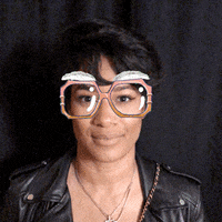 Video gif. A woman looks at us and pops her hands out in front of her as she says, “boo!” She wears a camera filter that gives her big madea glasses and eyebrows. 