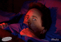 Seinfeld gif. Jerry Seinfeld as himself lies terrified in bed as he holds the blankets up tight to his face. He glances worried around the room.