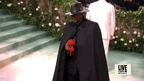 Met Gala 2024 gif. Usher, wearing an all-black custom Alexander McQueen ensemble, holds and lifts a red paper flower.