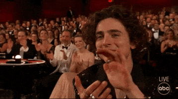 Celebrity gif. Timothée Chalamet sits in the audience at the 2022 Oscars, smiling and clapping. 