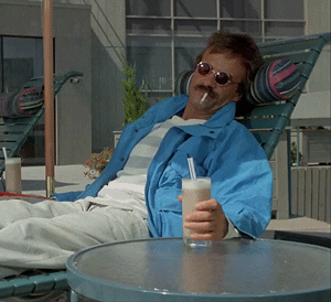 Movie Gif. Bruce Paul Barbour as the Beach Bum in Weekend at Bernie’s wears sunglasses and has a cigarette in his mouth as he sleeps in a beach lounge chair. He has a string around his wrist and someone pulls at it to wave his hand limply around.