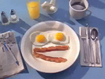 Video gif. A breakfast plate resembles a chattering face, with two over-easy eggs and two strips of bacon, and a hand comes in to dab toast in the yolk.