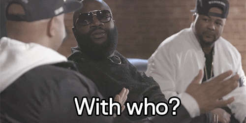 Celebrity gif. Rick Ross looks over at another man shrugging his shoulders and smiling as he says, “With who?”