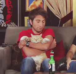 Video gif. Man holds a fat orange cat on his lap and embraces it. The cat sits on the man’s lap on its butt and has his arms stretched out as the man softly shakes and hugs the cat. The man rests his head on the cat’s head.