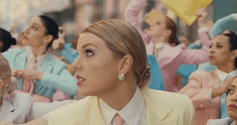 Music video gif. Taylor Swift in the Me music video is in a crowd of pastel business women while wearing a soft yellow suit. She turns to look straight at us and winks while giving a smug smirk. 