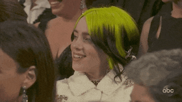 Celebrity gif. Billie Eilish sits in the audience of the Academy Awards. She looks up next to her, and cringes in embarrassment while smiling.