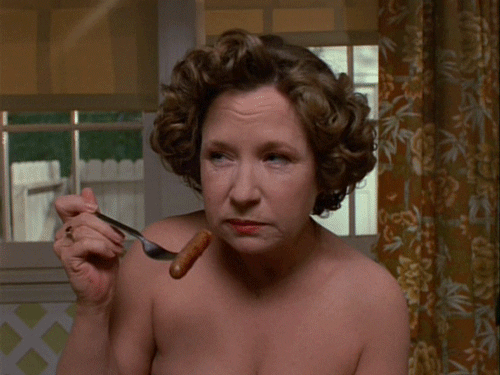 TV gif. Debra Jo Rupp as Kitty on That 70s Show, nude, stares suspiciously off to the side while chewing and holding up a breakfast sausage with a fork.