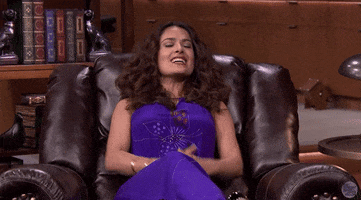 Tonight Show gif. Salma Hayek sits back in a large, leather armchair facing us as she smiles and lifts her fists above her head, shaking them in triumph. 