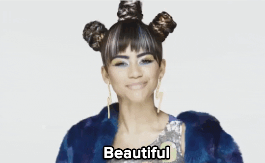 Celebrity gif. Zendaya with long dangling earrings, her hair in the shape of a crown with three buns, wraps herself in a blue fuzzy jacket, smiling and saying "beautiful," which appears as text.