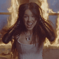 Music Video gif. Olivia Rodrigo in the Good Four U music video stands in front of a burning window as she screams at us. Her black leather gloved hands run down her face like she’s emulating the famous scream painting.