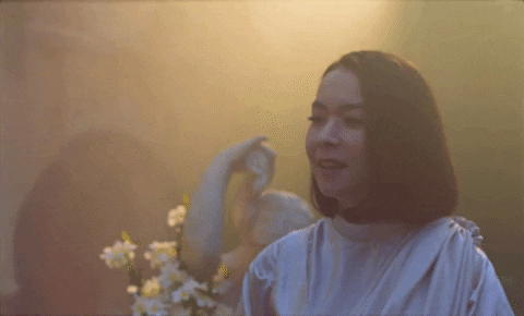 Music video gif. Mitski in her video for Stay Soft smiles blissfully as she steps toward us in a garden with golden light.