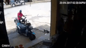 Video gif. A man on a motorcycle attempts to park his bike near a ledge, then tries to dismount but loses his balance. He falls over the ledge, his motorcycle falling on top of him. A crowd of people comes running to help him.