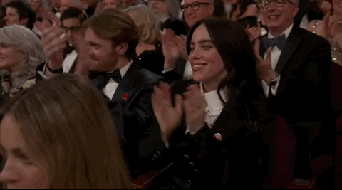 Oscars 2024 GIF. Billie Eilish, seated at the Oscars, applauds energetically, throwing in a whoop and cheer.