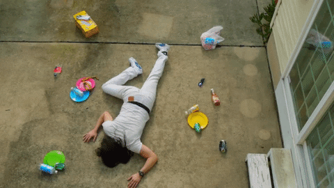 Music video gif. From the video for "It's Time to Party," Andrew WK lies facedown on pavement, surrounded by a few empty bottles, cans, and plates.