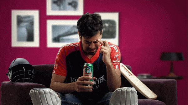 Sports gif. A man dressed in cricket gear and a Kingfisher jersey, beer in hand, sits on a couch, eyes closed and fingers crossed in anticipation. He looks up, shouts “no, no, no!” with hope, then scoffs, throwing his hand up in disgust and disappointment.