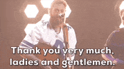 ACM Awards gif. Post Malone with a guitar wrapped around him motions closer to the standing microphone in a slighly goofy manner and says "Thank you very music ladies and gentlemen." 