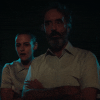 Celebrity gif. Kristen Stewart stands in the shadows behind David Cronenberg as they both gaze into the distance with their arms crossed.