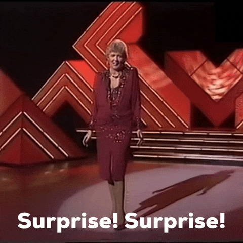 Video gif. A woman wearing a sparkling burgundy dress and jewelry stands underneath a spotlight on a stage with red and white light displays. She spreads her arms as she theatrically sings, "Surprise! Surprise!," which appears as text. 