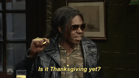 SNL gif. Dave Chappelle is wearing aviators and a leather jacket with his long hair tied in a ponytail. He smokes a cigarette and asks with a grin, "Is it Thanksgiving yet?"
