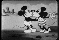 Cartoon gif. Mickey and Minnie are embracing and giving each other sweet kisses. They embrace and cozy up to one another as Minnie flutters her eyelashes at Mickey.
