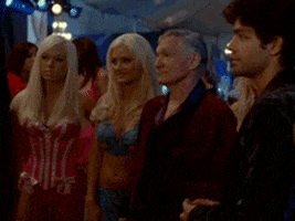 Celebrity gif. Hugh Hefner, surrounded by blonde Playmates dressed in lingerie, bobs his head to music with satisfaction.