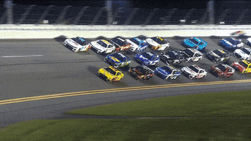 Video gif. Dozens of NASCAR race cars speed around the corner of a track clustered closely together in three long lines. 