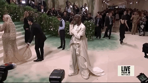 Met Gala 2024 gif. Lil Nas X poses on the red carpet holding up and flaring out his long spiky nails with diamond studs on clear polish. He's wearing a monochrome ivory-colored Luar suit with a full trench coat that falls and drapes slightly along the floor. His outfit is accented by irregular Swarovski sequin panels.