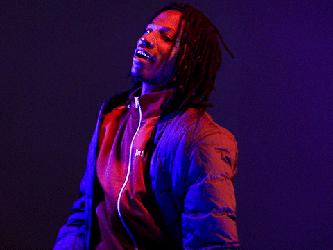 Celebrity gif. Joey Badass wears an unzipped puffer jacket with a cool smile, lit by red and blue lights and shimmying his shoulders.