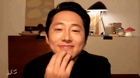 Celebrity gif. Steven Yeun is on webcam and has his hand on his chin. He smiles and leans back before waving at us.